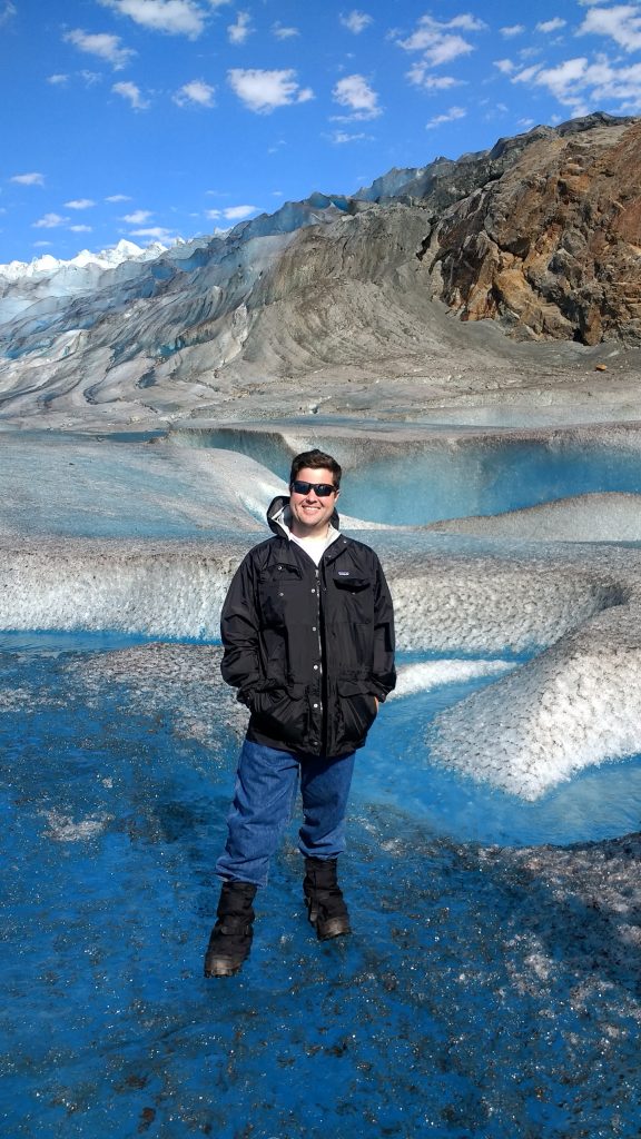 Full shot of a smiling man wearing sunglasses and a black winter jacket standing on vibrant blue and white glacier ice under a bright blue sky.