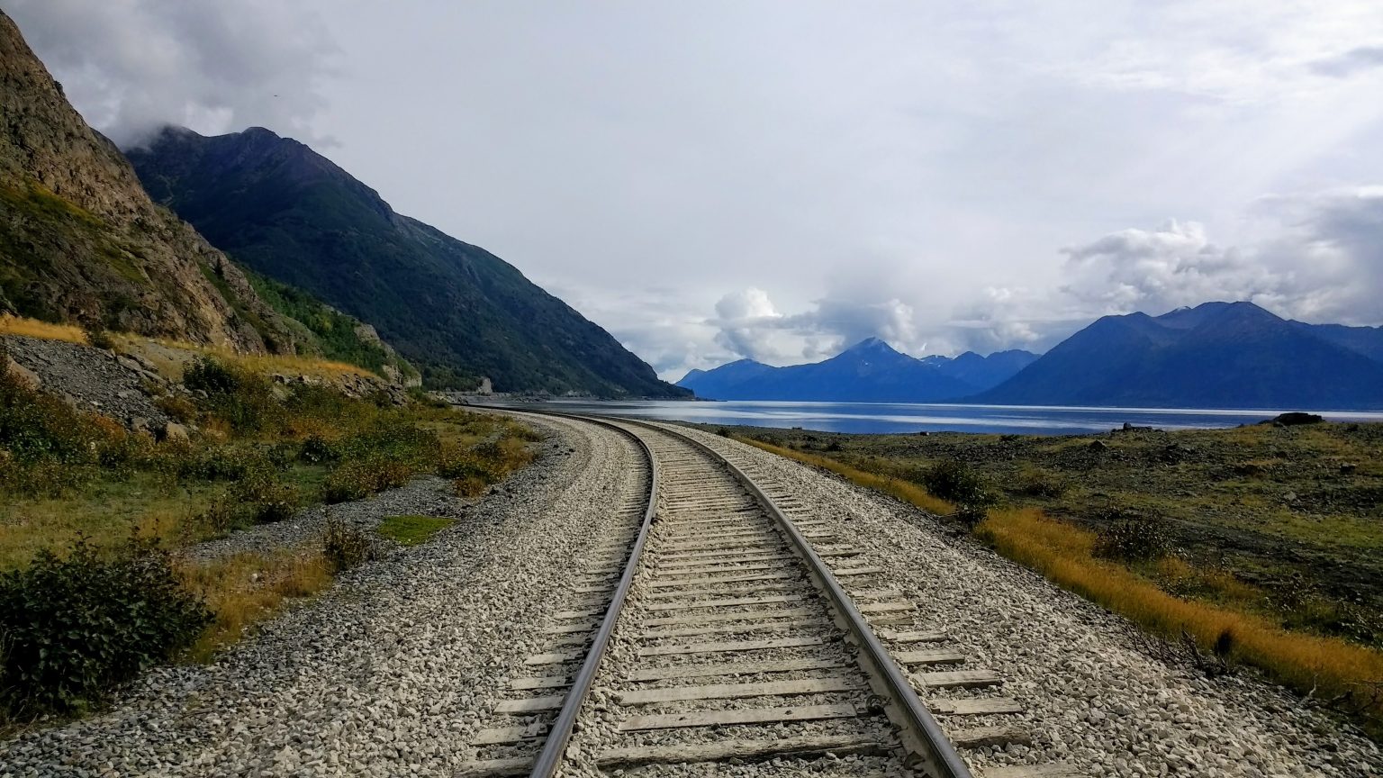 A scenic perspective shot following train tracks as they curve alongside a vast lake bordered by massive, dark mountains under a bright cloudy sky.