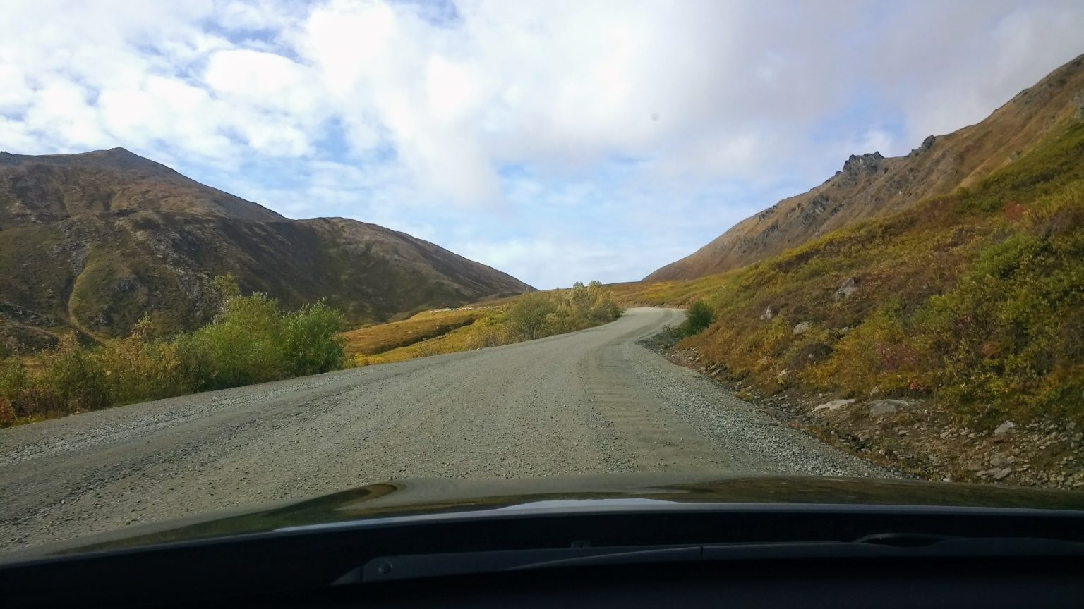 A wide-angle landscape shot from inside a vehicle looking out at a winding gravel road heading through rolling green and rocky hills under a cloudy sky.